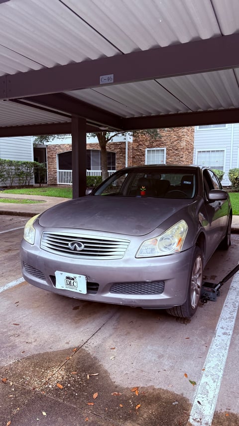 Silver Infiniti sedan parked under a metal carport with brick house and green lawn in the background