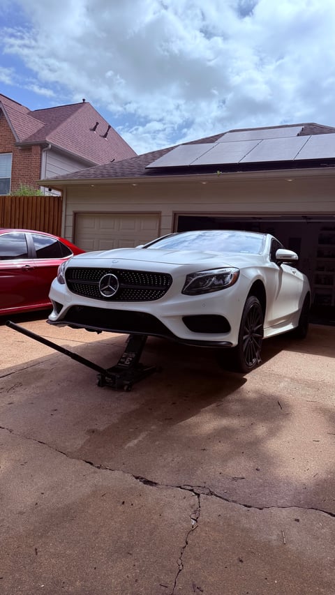 White Mercedes-Benz coupe parked in a driveway next to a brick home with solar panels and an open garage