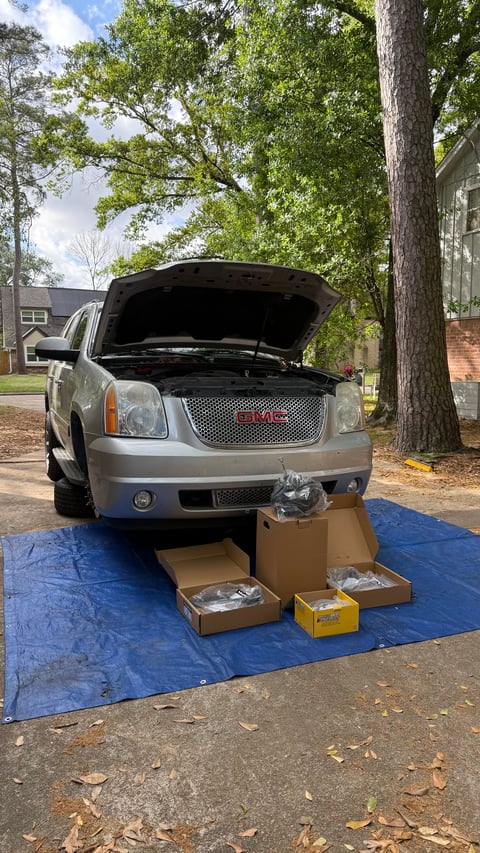 Silver GMC Yukon SUV with open hood and engine removed, displayed on blue tarp with cardboard boxes and engine parts