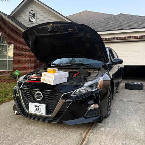 Black Nissan sedan parked in driveway with open hood displaying engine parts and boxes, brick house in background