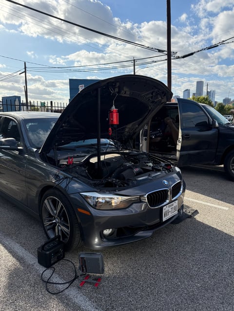 BMW sedan with open hood displaying engine, connected to portable jump starter in parking lot with cityscape background