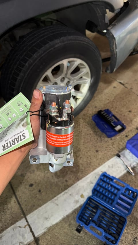 Person holding a car starter motor in front of a vehicle tire, with tools and manuals visible on the ground